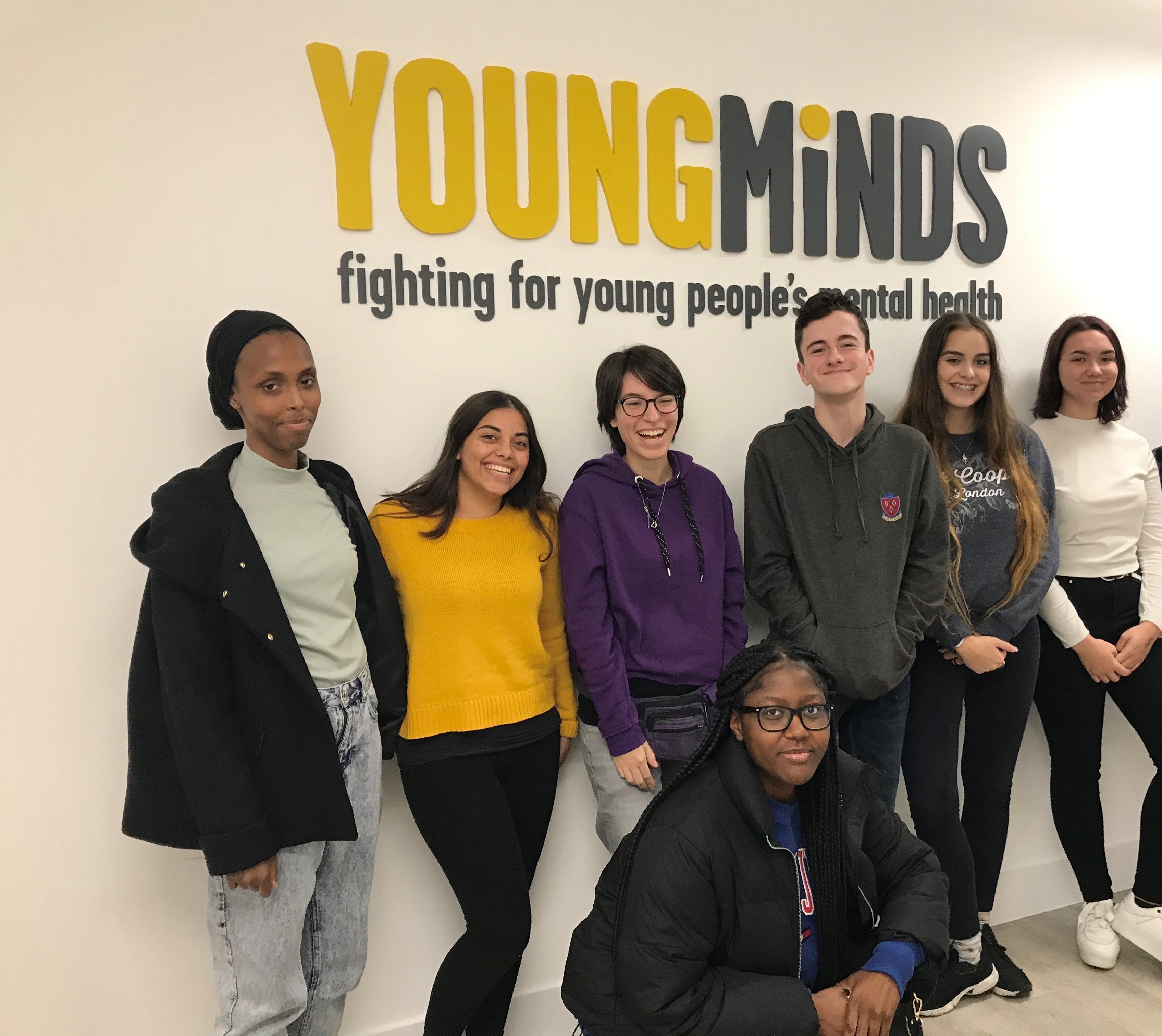 Our Youth Panel stand against a white wall with our logo in yellow and grey. The youth panel are standing together smiling and looking at the camera. Underneath the logo it reads: fighting for young people's mental health.