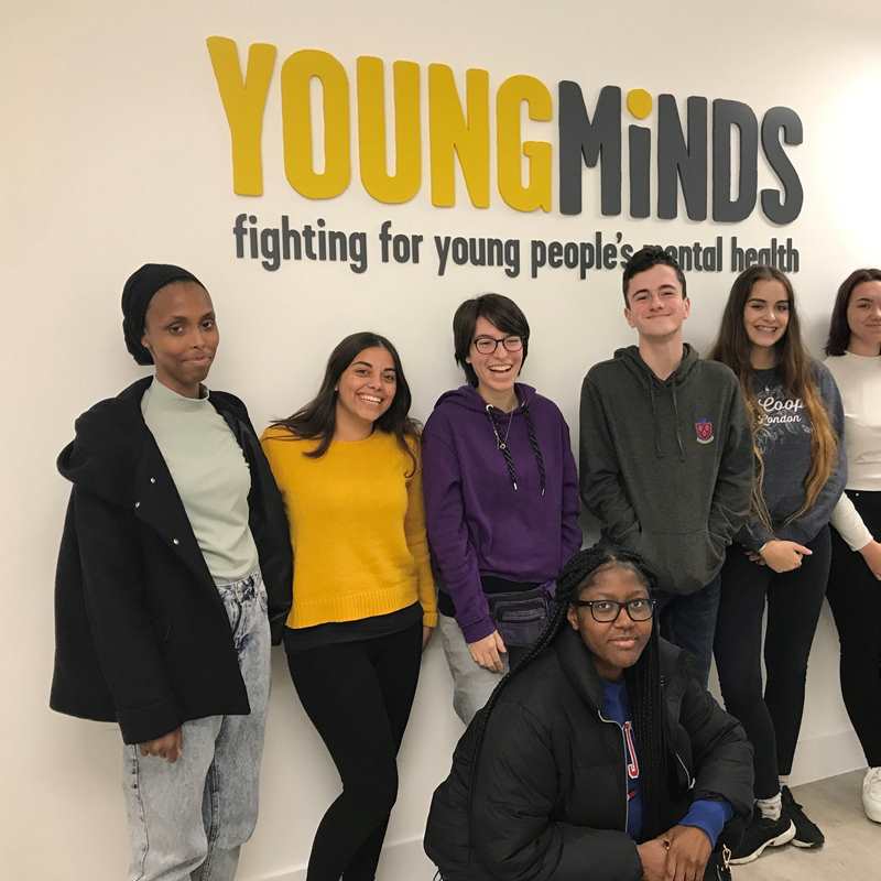 Our Youth Panel stand against a white wall with our logo in yellow and grey. The youth panel are standing together smiling and looking at the camera. Underneath the logo it reads: fighting for young people's mental health.