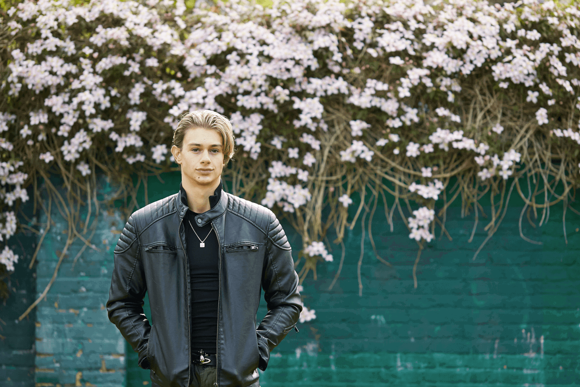a boy wearing black jacket with hands on his pockets standing and looking in front of the camera on the backgound is a wall with flowers