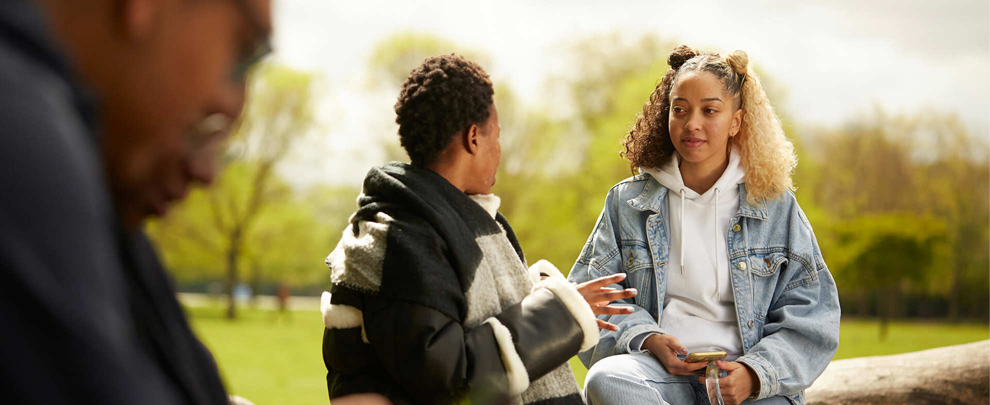 Two girls sit and chat on a fallen tree trunk in a park.