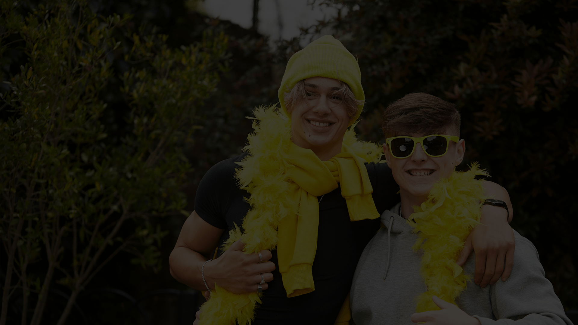 Two young people with their arms around each other. They are wearing a yellow feather boa, yellow hat and yellow sunglasses.